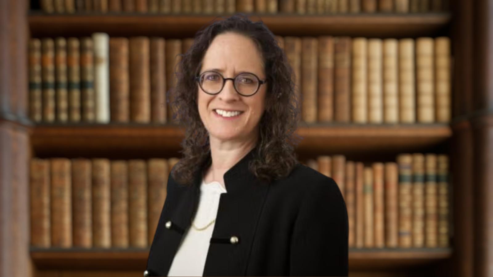 Alyza Lewin, glasses, black jacket, in front of bookshelf bg